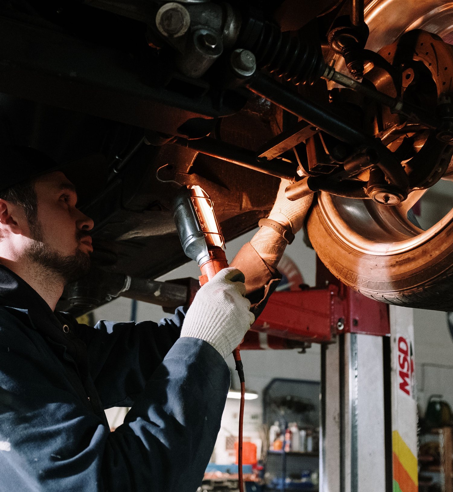 A mechanic running an impact wrench on a wheel hub under a vehicle raised on a two-post lift.